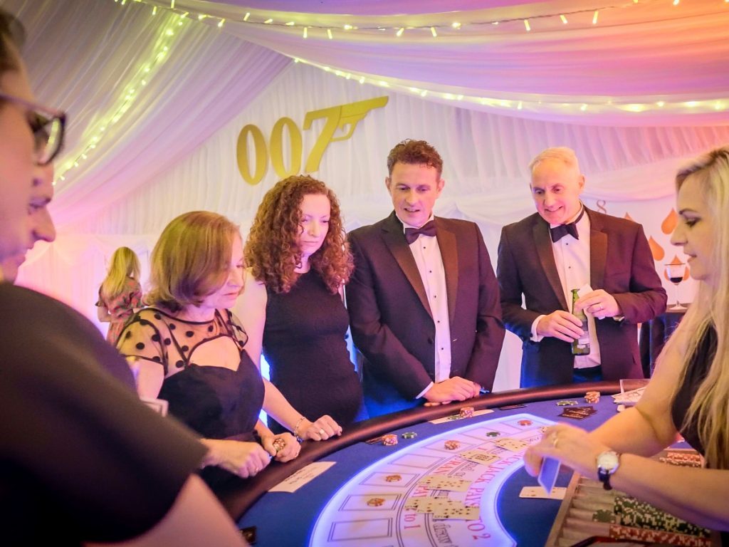 Various people dressed in black tie around a gambling table within a marquee decorated with a James bond Theme